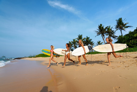 Friends walk on tropical beach holding surfboards. Young surfers explore ocean waves under blue sky. Adventure surf camp. Summer travel, beach sports. Female surfers enjoy sun, sand in paradise.の写真素材