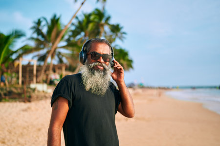 Senior stylish bearded man enjoys music on sunny beach. Modern grandpa with sunglasses and headphones embraces tech and freedom. Active lifestyle and youthful spirit by ocean.の写真素材