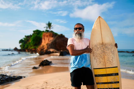 Senior man with beard holds surfboard on sunny beach. Elderly surfer smiles, enjoys active retirement on ocean shore. Happy mature male embraces vibrant lifestyle. Summer adventure, freedom.の写真素材