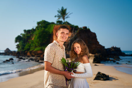 Romantic couple stands on tropical beach at sunset, man holds bouquet, embracing woman. Smiling lovers enjoy Valentines Day, posing for memory. Happiness, love, travel concept.の写真素材