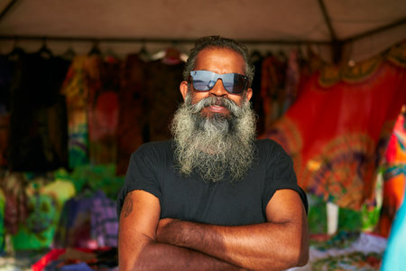 Senior artist with big beard, sunglasses stands in front of colorful tie-dye t-shirts at vibrant...