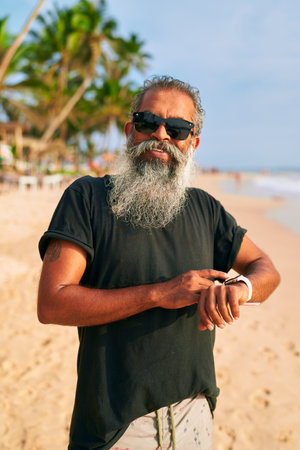 Senior bearded man with sunglasses on sandy beach uses smart watch. Modern grandpa enjoys tech, staying fit by the sea. Active old stylish male uses wearable device in sunny tropical location.の写真素材