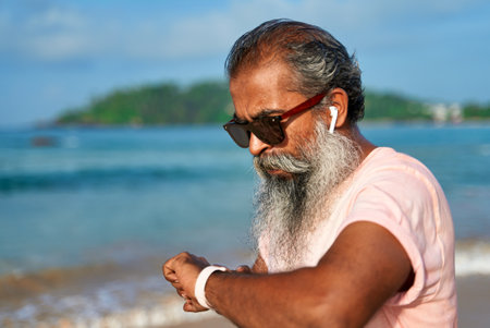 Senior bearded man checks smart watch on beach while jogging. Old male wears wireless earbuds....
