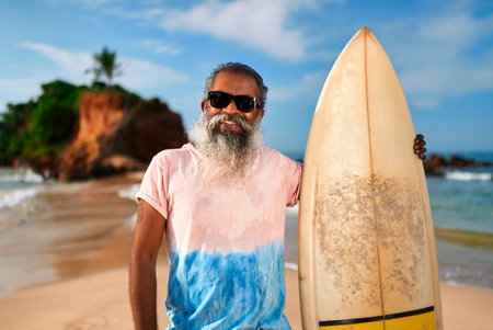 Senior bearded man with surfboard on sunny beach. Elderly surfer enjoys active lifestyle by ocean. Older male in sunglasses embraces sea adventures and fitness. Travel, leisure vibes.の写真素材