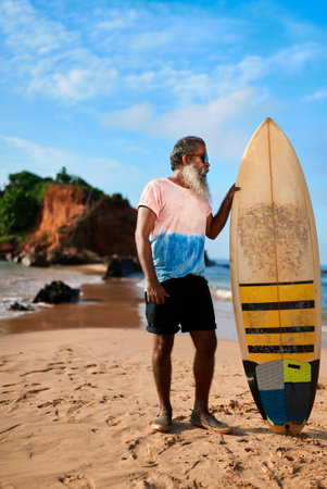 Senior man with surfboard stands on sandy beach. Elderly surfer poses under blue sky by rocky coast. Active retiree enjoys outdoor sea adventure. Tropical surf lifestyle travel concept.の写真素材