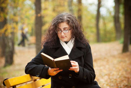 young woman reading book in autumn park benchの写真素材