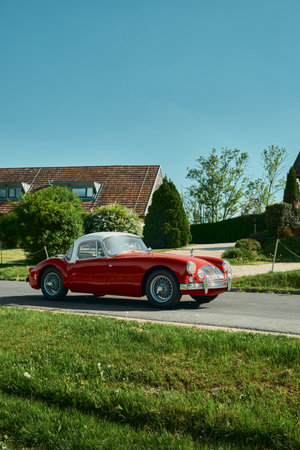 Red retro car driving on a road with neatly trimmed grass in the foreground, a building with a red roof in the background, and a clear blue sky, capturing a charming countrysideの写真素材