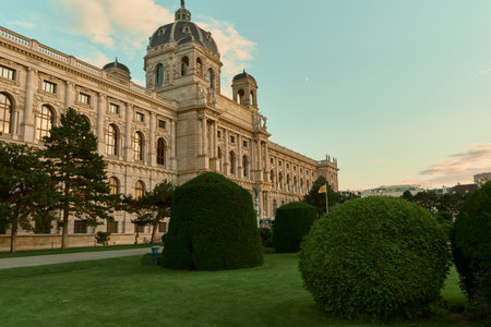 View of Vienna's Kunsthistorisches Museum with pine trees, manicured garden shrubs, blue sunset sky and a visible moon, combining nature and historic architecture.の写真素材