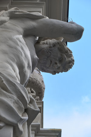 Detailed low-angle view of a muscular white marble or plaster male figure, possibly an Atlas, supporting weight on his head, with blue sky and architectural relief in the backgrounの写真素材