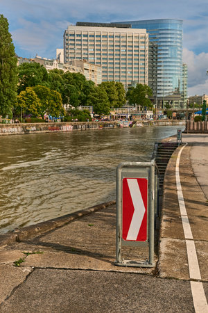 Urban riverside scene featuring red and white turn sign on pavement, concrete steps leading to water, and modern buildings with trees across the river.の写真素材
