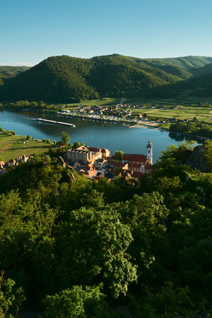 Vertical sunny view from above of a baroque church, historic town, forests, vineyards, hills, and the Danube River with a passing barge in Wachau Valley, Austria.の写真素材