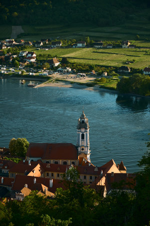 Vertical sunny view highlighting a baroque church, red-roofed houses, wide Danube River, and surrounding vineyards in Wachau Valley, Austria. Scenic and vibrant landscape.の写真素材