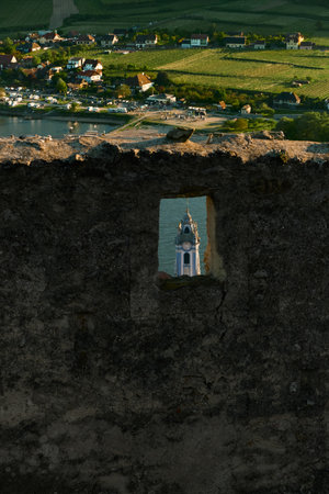 Scenic view through stone window of ruined castle, focusing on blue baroque church with sunlit vineyards in background on a bright summer day in Austria.の写真素材