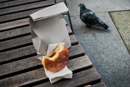 Bitten donut resting on a napkin next to a paper bag on a wooden bench, with a pigeon walking...