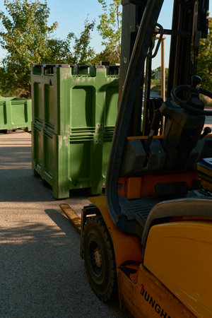 Vertical photo of a yellow loader with green crates filled with grapes on a vineyard road. Sunlit...