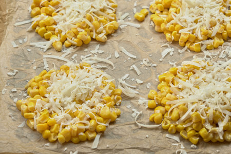 Close-up top-down view of corn rounds on parchment paper, topped with grated cheese, ready for...