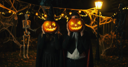 A woman dressed as a witch and a man as Dracula hold glowing pumpkins at head level in a Halloween-decorated street with lights, skeletons, garlands, and cobwebs.の素材