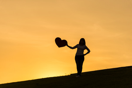 Silhouette of asian lady standing on hill and holding heart with one hand on the side during sunset time to show her love on Valentines dayの写真素材