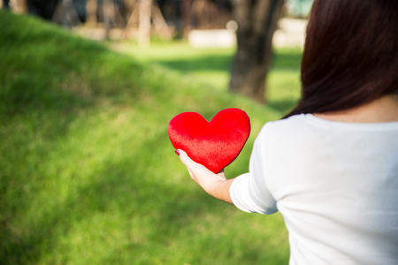 Asian woman holding red soft heart pillow with left hand at park as background to show her love on Valentines day concept with warm sun lightの写真素材