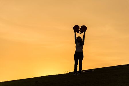 Silhouette of asian woman standing on hill and holding broken heart with two hands during sunset time to show her love on Valentines day, selective focusの写真素材