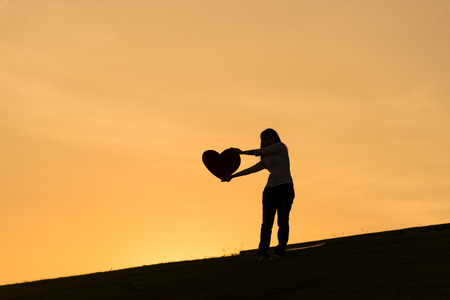 Silhouette of asian lady standing on hill and holding heart over her head with two hands during sunset time to show her love on Valentines day, selective focusの写真素材