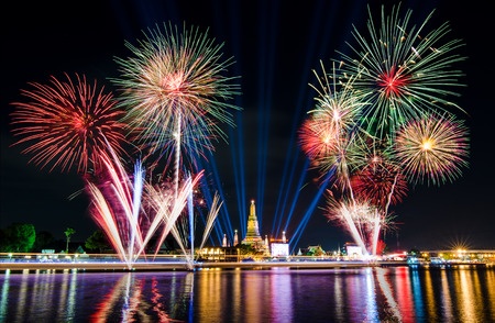 Fireworks lit up beautifully over dark sky and golden pagoda of temple close to the river with city light, blue beam laser and colorful reflection at night time to celebrate new year occasionの写真素材
