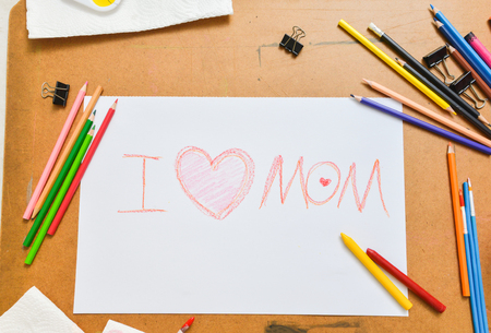 Sketch paper with a sentence "I love mom" surrounding by pencils, painting brush, tray of white, yellow, blue, red, green, yellow color and black clips over cork board in art class, top viewの写真素材