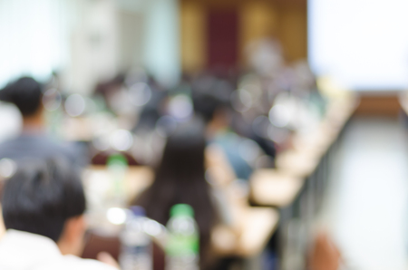 Blur background of people sitting in seminar or big meeting room with bokeh, chairs, tables and white slide screen to be used for business, training or seminar promotion / advertisingの写真素材