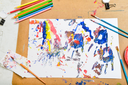 Poster color art on white sketch paper with pencils, painting brush, colour tray of white, yellow, blue, red, green, and black clips over cork board backgroundの写真素材