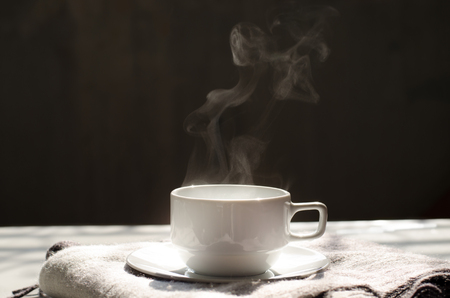 Hot black coffee in white ceramic cup and saucer on cozy wool scarf with nice steam warm sunlight in morning and copy space for text, wording insertion over dark grey background, hot beverage for winter conceptの写真素材