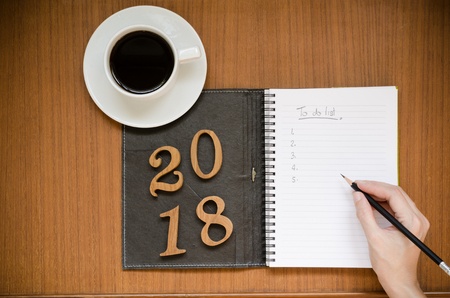Recycled notebook on blank page white paper and dark cover with 2018 wood letters & black sharp pencil over teak wood background with copy space and black coffee in white cup and saucer, office or education conceptの写真素材