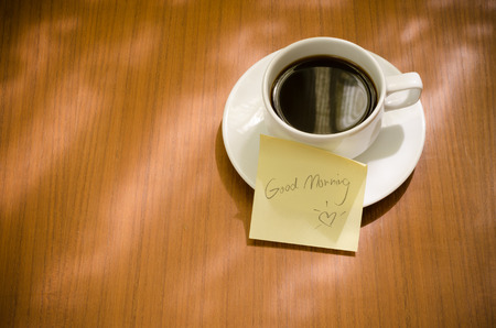 Black coffee in white ceramic cup and saucer with Good Morning word on yellow sticky note paper and warm morning gold yellow sun light through window and shadow on wooden tableの写真素材