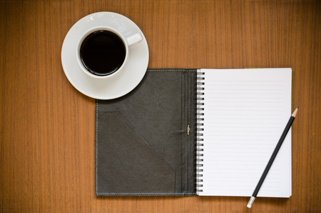 Recycled notebook on blank page white paper with lines and dark cover with black sharp pencil over teak wood background with copy space and black coffee in white cup and saucer, office or education conceptの写真素材