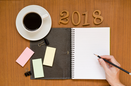 Recycled notebook with woman hand writing in the empty paper note surrounding with black coffee in white cup, 2018 letters, yellow, pink, green sticky pads, pencil & paper clips on dark wood background spaceの写真素材