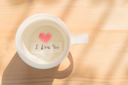 White coffee cup with paper cut in heart shape painted with red pencil and I love you in black on wood background under sunlight showing love relation concept on Valentines day or wedding celebrationの写真素材