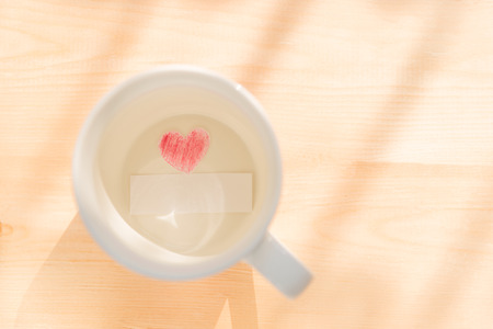 White coffee cup with paper cut in heart shape painted with red pencil and empty or blank paper card on wood background under sunlight to show love relationship on Valentines day or wedding partyの写真素材