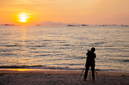 Silhouette of professional photographer shooting sunset with good lens, camera and tripod at the beach on sunny hot day in summer with calm clear sea and skyの写真素材