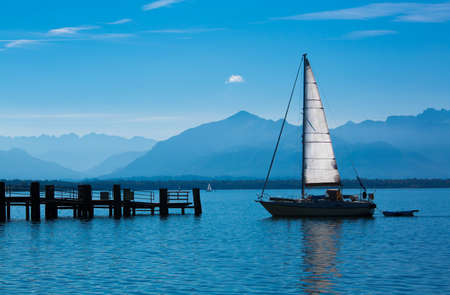 Sailboat on a quiet lake with a wooden pier and mountains in the backgroundの写真素材