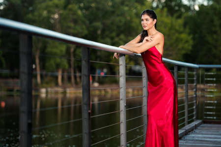 Beautiful young woman wearing a long red silk formal dress in the gardens in the afternoon.の写真素材