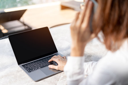 Rear view of young business woman talking on the phone while sitting at her workplace in front of laptop. Entrepreneur freelance working with laptop.の写真素材