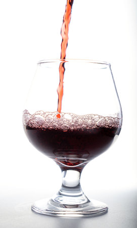 A stream of red liquid, wine or juice, being poured into a large clear glass with foam and bubbles against a bright white background.の写真素材