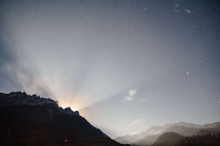 Moon rising with ray light behide the mountain with little snow on the top and moving cloud in the blue color night with stars in winter at Lachung in North Sikkim, India.の写真素材