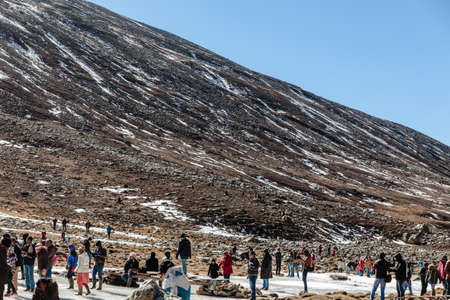 Black mountain witn snow and below with tourists on the ground with brown grass, snow and frozen pond in winter at Zero Point at Lachung. North Sikkim, India.のeditorial素材