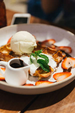 Chocolate waffle topping with scoop of vanilla and strawberry ice cream, whip cream, sliced strawberry, mint leaves, icing and chocolate sauce on blue plate at dessert cafe in Bangkok, Thailand.の写真素材