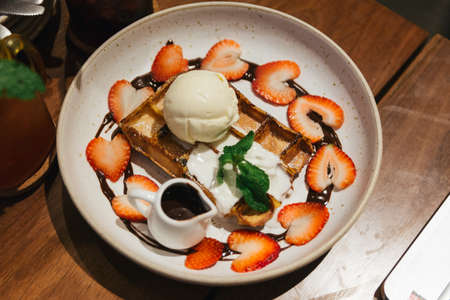 Chocolate waffle topping with scoop of vanilla and strawberry ice cream, whip cream, sliced strawberry, mint leaves, icing and chocolate sauce on blue plate at dessert cafe in Bangkok, Thailand.の写真素材
