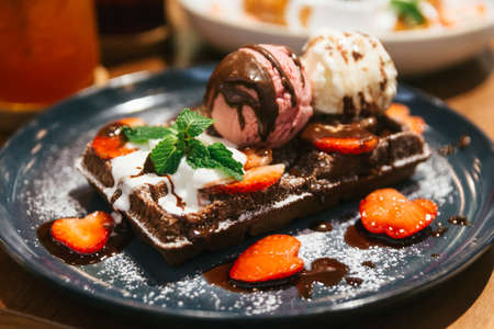 Chocolate waffle topping with scoop of vanilla and strawberry ice cream, whip cream, sliced strawberry, mint leaves, icing and chocolate sauce on blue plate at dessert cafe in Bangkok, Thailand.の写真素材