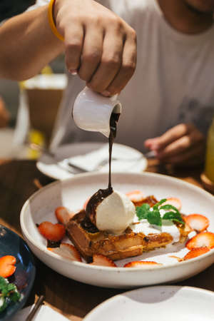 Pouring Chocolate sauce on waffle that topping with a scoop of vanilla ice cream, whip cream, sliced strawberry and mint leaves on white plate at dessert cafe in Bangkok, Thailand.の写真素材