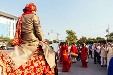Indian groom ridding white horse with yellow and red pattern fabric, flower necklace and red turban...