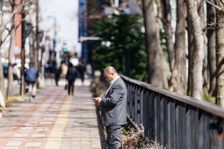 Business man looking on his smart phone on the street walk way with blur background in Sapporo at Hokkaido, Japan.