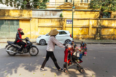 Hanoi street with a man driving motorcycle and woman with mobile retail cart and moving car in Vietnam.のeditorial素材